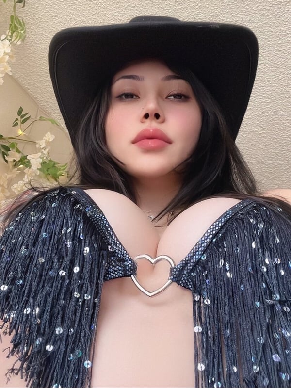 A person wearing a black cowboy hat and fringed top, posing with plants in the background.