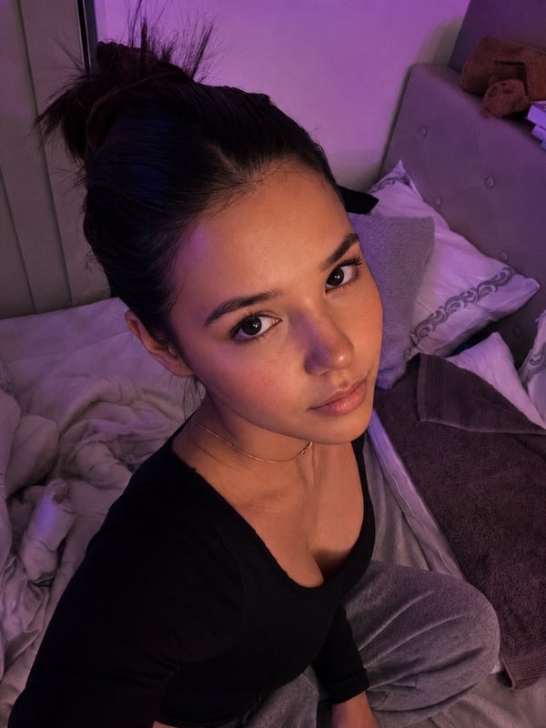 Young woman with long dark hair lying on a pink fluffy rug in a bedroom with pink lighting, holding a phone for a mirror selfie.