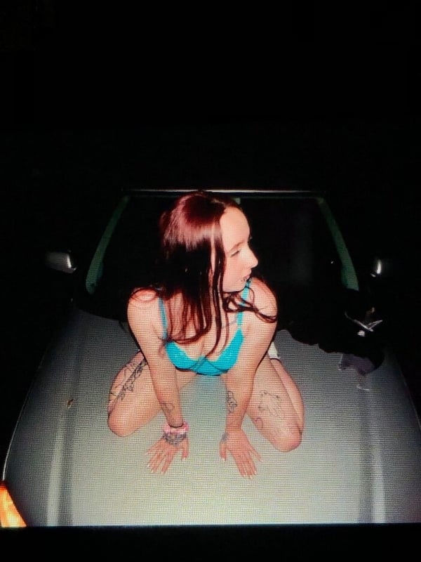 A woman with reddish hair sitting on a car hood at night, dressed in a blue swimsuit, showing tattoos on her arms.