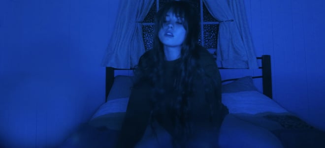 A young woman with long dark hair sits on a bed in a dimly lit room with blue tones and draped curtains behind her.