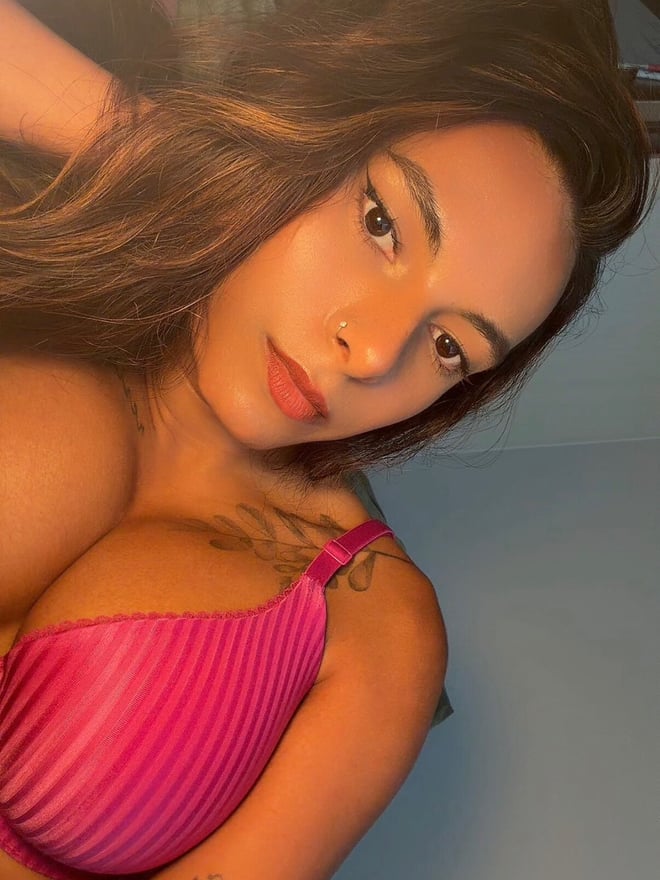 Close-up portrait of a woman with wavy brown hair wearing a pink top, showing tattoos on her shoulder.