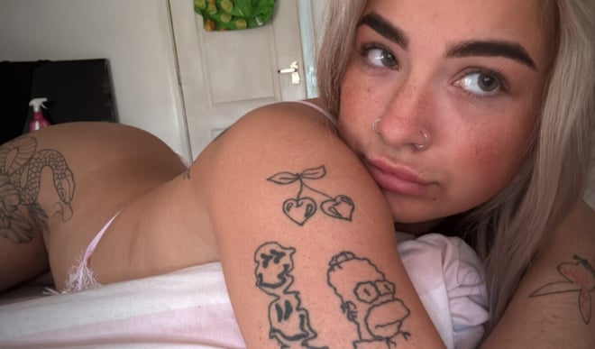 A young woman with blonde hair lying on a bed in lingerie, surrounded by cartoon drawings and houseplants in a cozy bedroom setting.