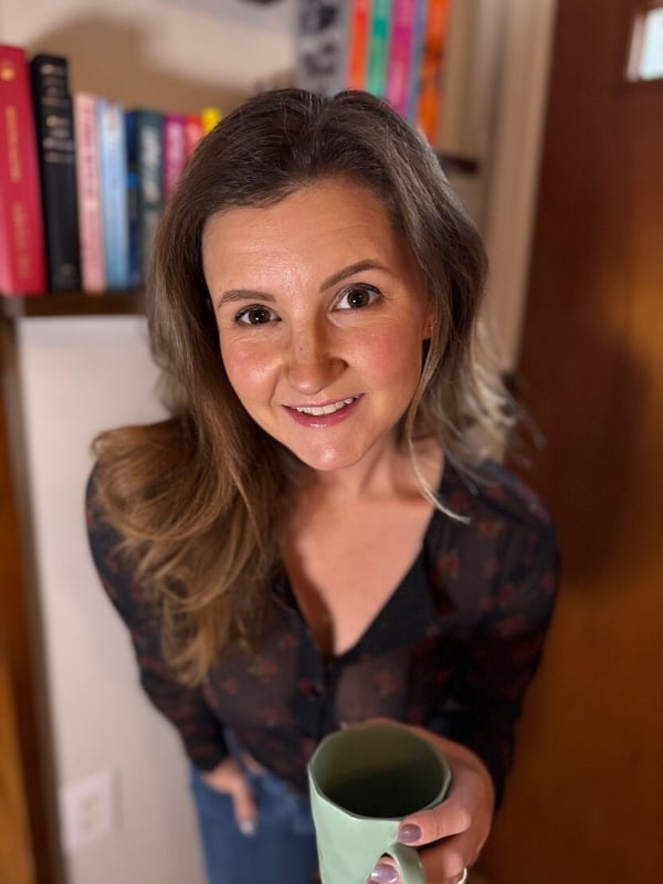 Woman with brunette hair smiling while holding a green mug, standing in front of bookshelves in a cozy indoor setting.