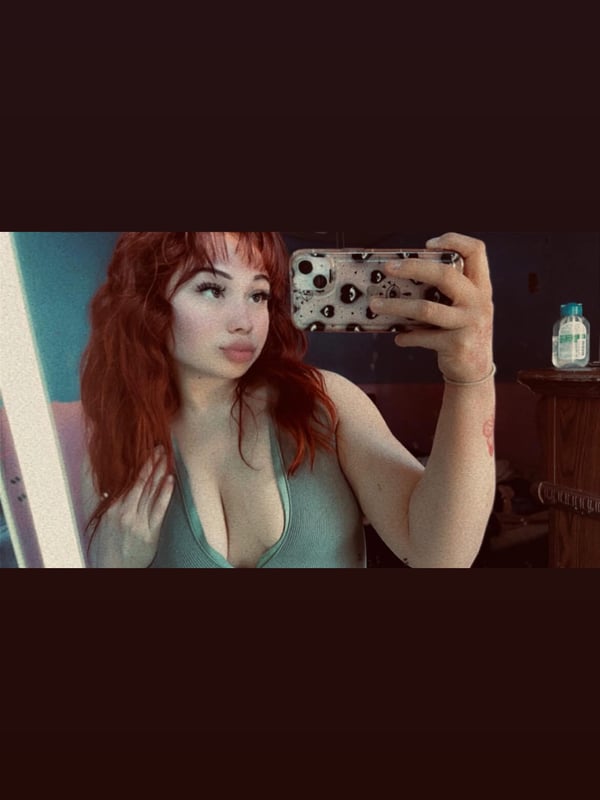 A young woman with red hair taking a mirror selfie in a low-cut teal top, holding her phone with a spotted case in a bathroom setting.