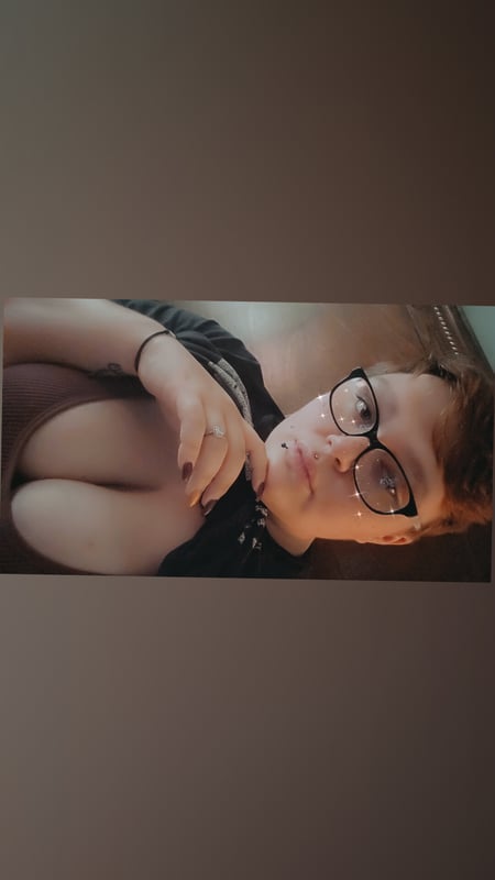 A woman with short hair and glasses lying on her side, wearing a dark top, posing thoughtfully indoors.