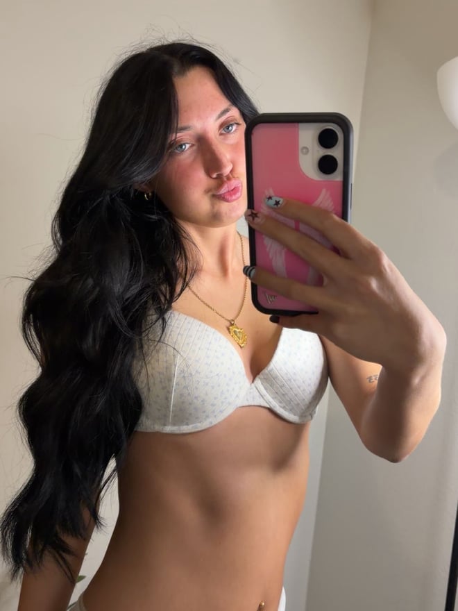 Young woman with wavy dark hair and blue eyes smiling at the camera in a bedroom, wearing a gray tank top and gold necklace.