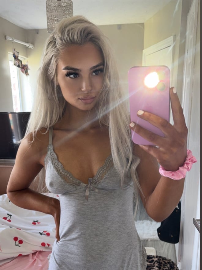 Young woman with platinum blonde hair taking a selfie in a bedroom, wearing a gray lace-trimmed slip dress and pink wristband, smiling softly.