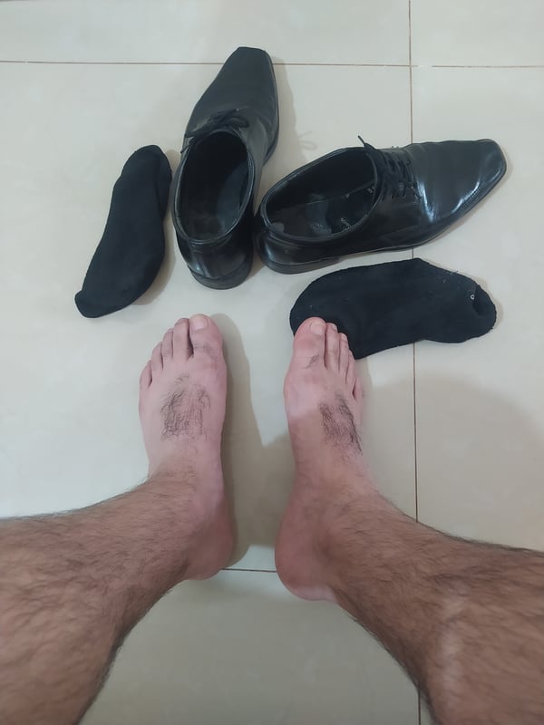 Close-up view of bare male feet with visible hair on legs and toes, positioned next to black socks and dress shoes on a tiled floor.