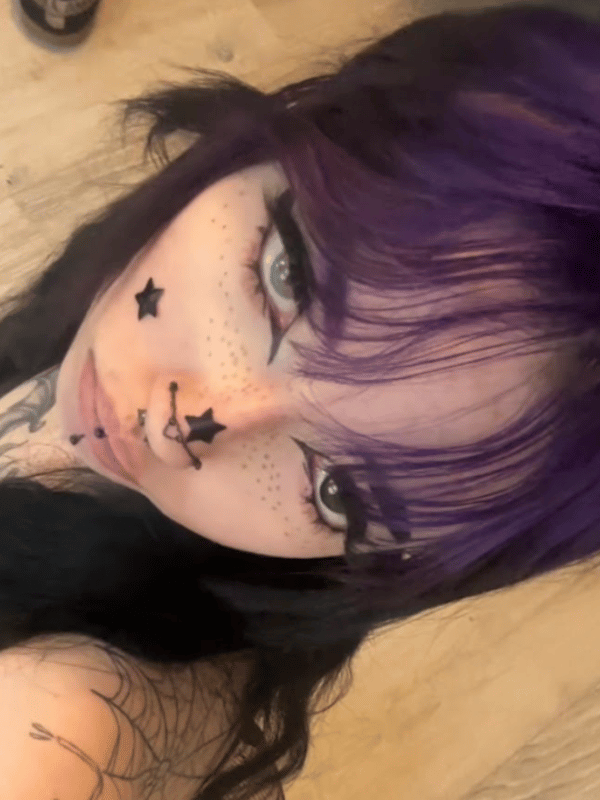 A close-up selfie of a person with purple hair and face tattoos looking at the camera.
