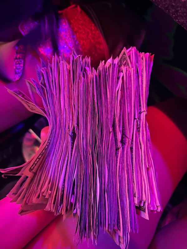 Stacks of cash arranged in a pile under vibrant purple and pink lighting with subtle background elements.