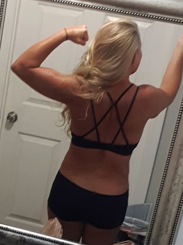 A woman with long blonde hair flexing her arms in a black sports bra and shorts, posing in front of a mirror in a bedroom.