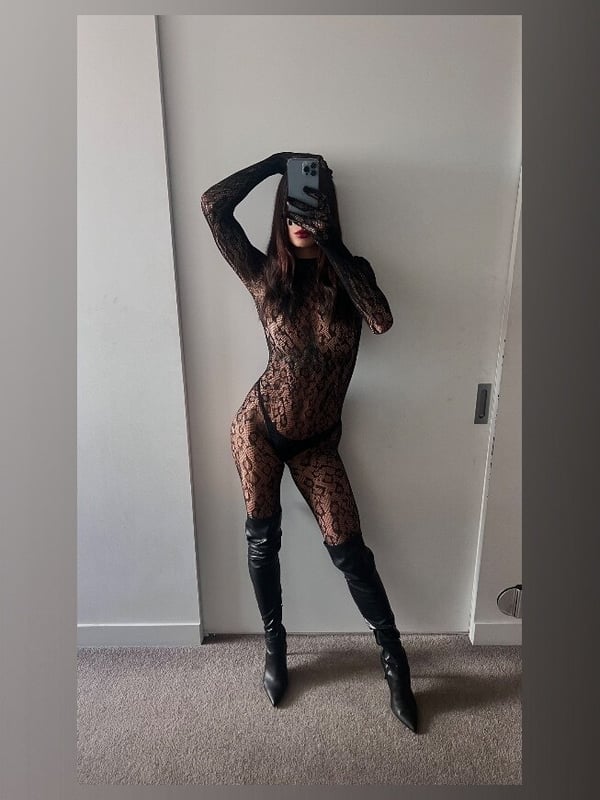 A woman striking a pose in a form-fitting outfit, long boots, and a face mask against a plain wall.