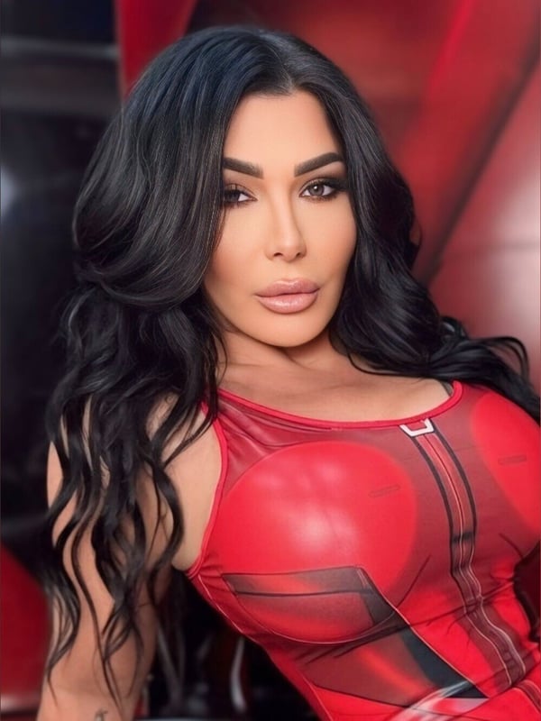 Close-up portrait of a person with long dark hair, striking makeup, and a red mesh top, smiling seductively with a red background.