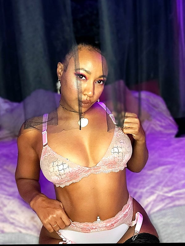 A woman sitting on a bed in sheer pink lingerie with purple lighting and sheer curtains in the background.
