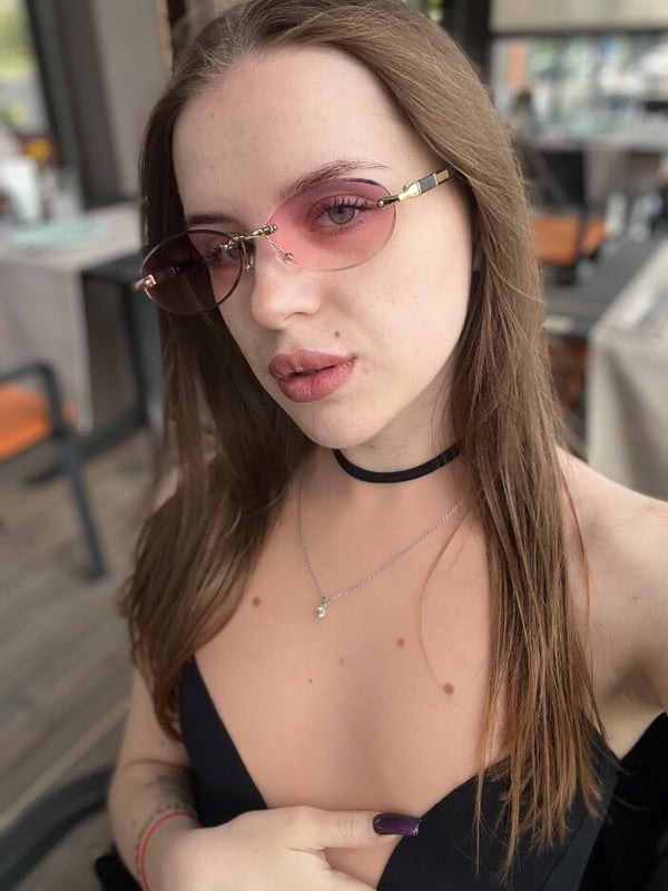 Young woman with long brown hair wearing sunglasses and a choker necklace, posing indoors near a window.
