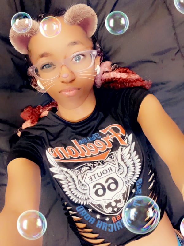 Young woman with puffy hair buns and glasses, lying on a bed surrounded by bubbles, wearing a cropped graphic t-shirt and low-rise bottoms.