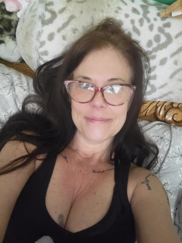 Woman lying on a bed with plush toys and pillows, wearing a black tank top and glasses, smiling at the camera.