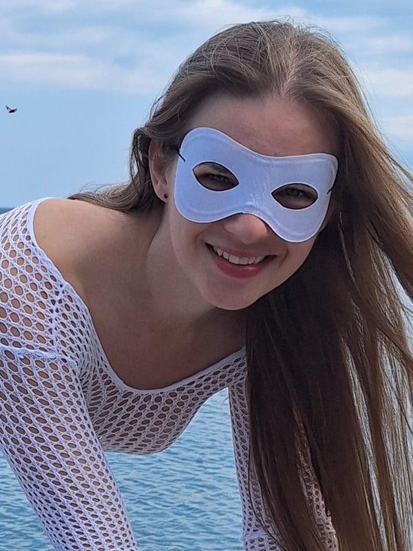 A smiling woman with long brown hair and a white mask poses in a sheer white top against a beach background.