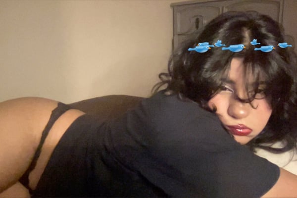 A young woman with long dark hair and a blue flower crown lies on her side on a bed, wearing a black top and bottoms, in a softly lit bedroom with wooden furniture.