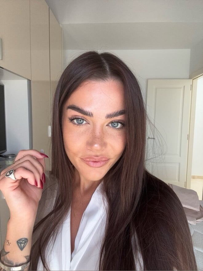 A woman with long straight brown hair and blue eyes smiling at the camera, wearing a white robe in a modern bedroom setting.