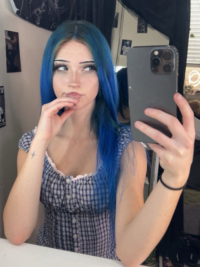 A young woman with long blue hair posing for a mirror selfie in a checked top.