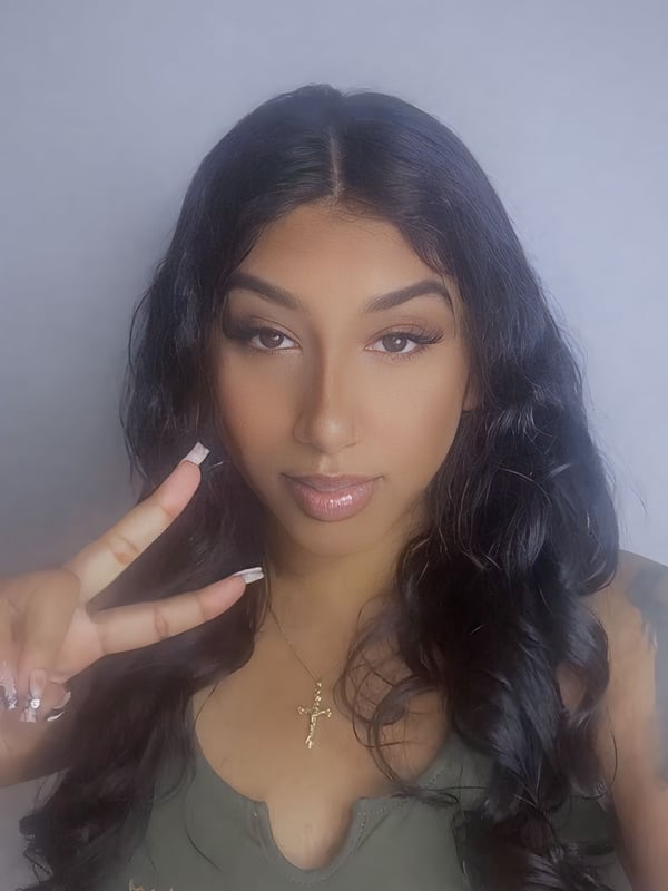 Young woman with long dark hair making a peace sign, wearing a green v-neck top and jewelry, posing against a light background.