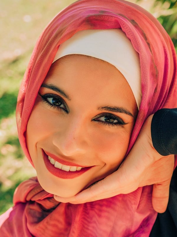 A smiling woman wearing a pink hijab and white headscarf, posing with her hand on her cheek against a green outdoor background.