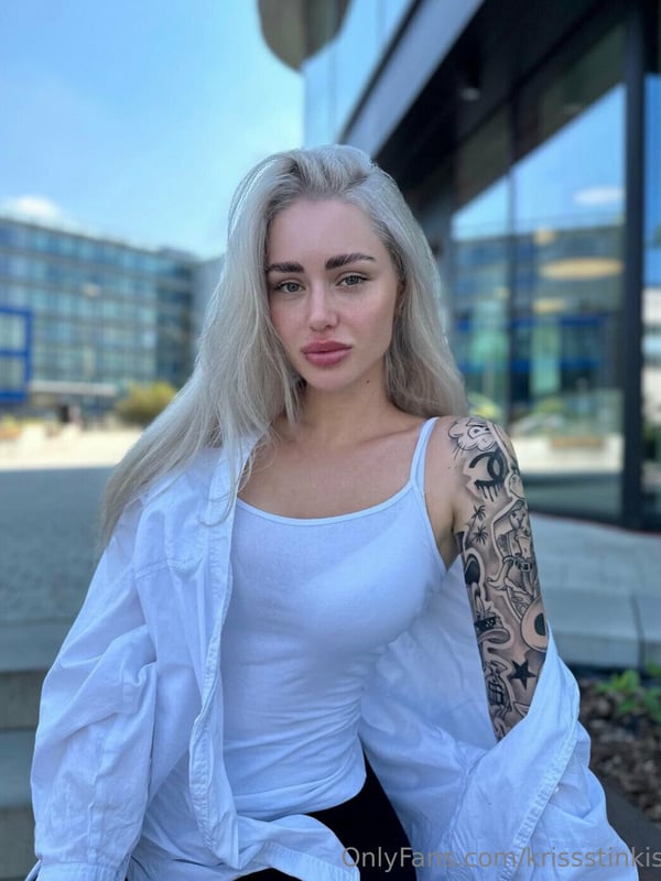 A woman with platinum blonde hair standing outdoors in front of modern buildings, wearing a white tank top and displaying tattoos on her arm.