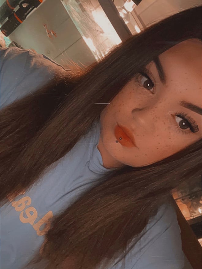 A young woman with long brown hair smiling at the camera, wearing a light blue hoodie with 'baby' text, in a home setting with warm lighting.