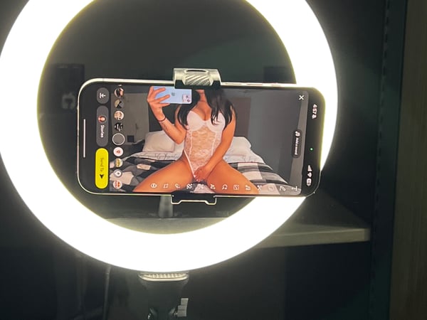 A person sitting on a bed in lingerie, holding a smartphone with their reflection visible in the screen, positioned in front of a ring light.