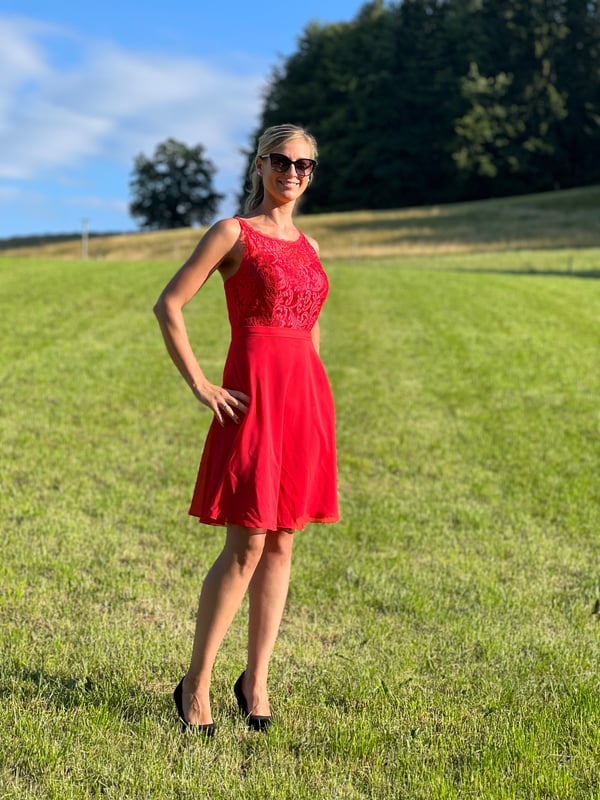 A woman standing in a green field wearing a red lace dress and black high heels, with sunglasses and blonde hair, against a sunny forest background.