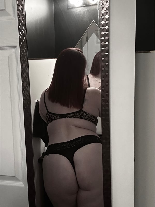 A woman standing in front of a mirror in a bathroom, wearing black lingerie, viewed from behind.