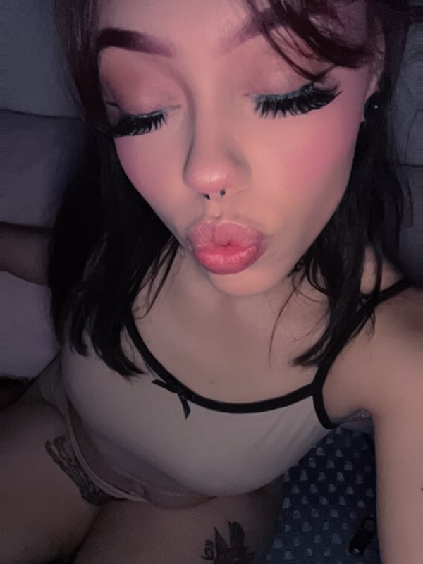 Close-up portrait of a young woman with long dark hair, heavy makeup, and a playful pout, wearing a camisole top.
