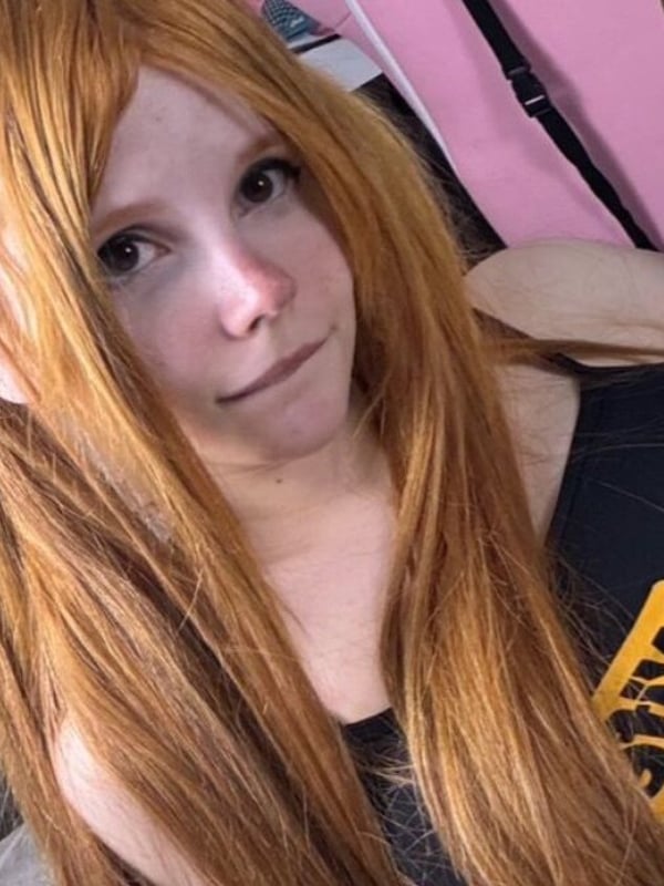 A close-up selfie of a woman with long orange hair in pigtails wearing a black tank top.