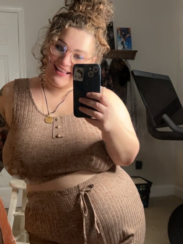 A smiling woman with curly hair and glasses taking a selfie in a cozy home gym setting, wearing a knit top and pants.