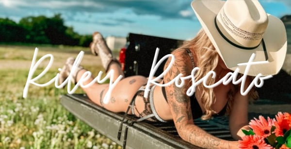 A woman in a cowboy hat and black bikini posing playfully on a truck bed in a green field with wildflowers and blue sky.