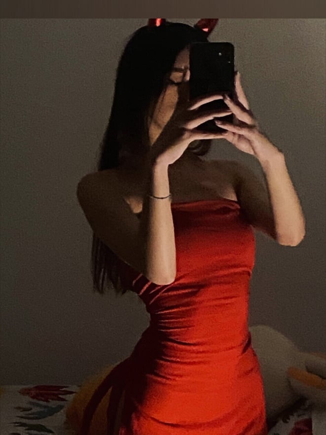 A young woman with long dark hair posing indoors beside a bed and side table.