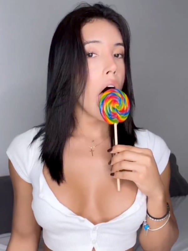 A person with long dark hair wearing a white top, holding and licking a large colorful lollipop against a gray background.
