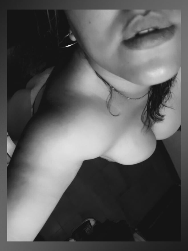 Close-up black and white portrait of a woman with wavy hair, earrings, and necklace, posing with lips pursed against a dark background.