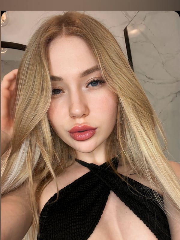 A young woman with long blonde hair smiling at the camera, wearing a black halter top, with soft lighting and mirrors in the background.