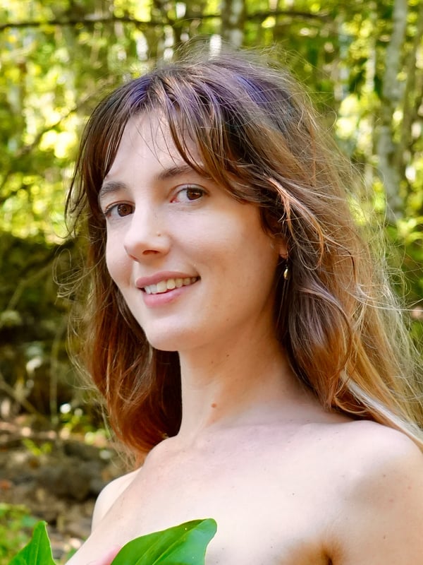 A smiling woman standing in a verdant forest, holding a large green leaf against her chest amid dense greenery.