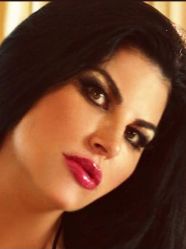 Close-up portrait of a woman with long dark hair, bold makeup, and glossy lips, gazing at the camera.