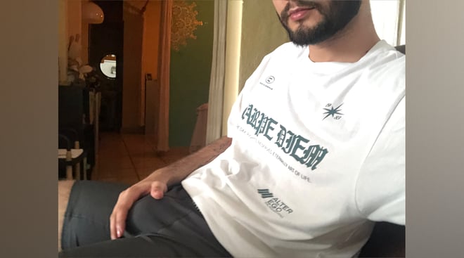 A man sitting casually in a white graphic t-shirt and dark pants inside a warmly lit room with green walls and furniture.