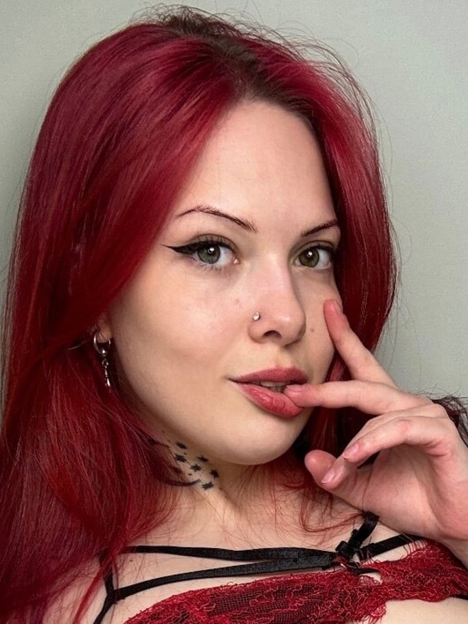 Close-up portrait of a young woman with vibrant red hair, green eyes, and a nose piercing, wearing lace lingerie and making a playful gesture with her finger on her lips.