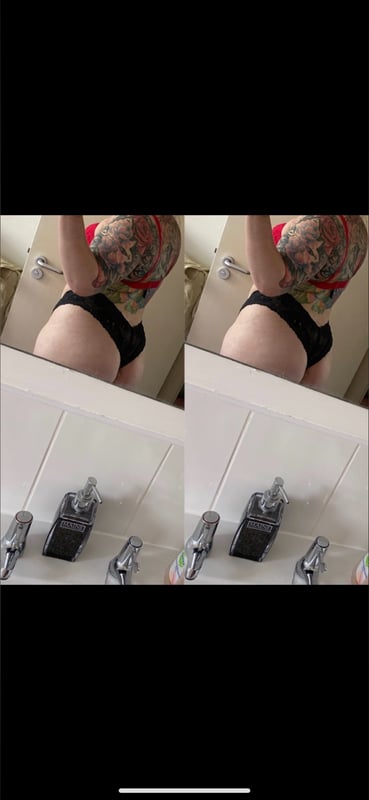 Woman standing in a bathroom mirror, wearing black underwear and showing tattoos on her arms and back.