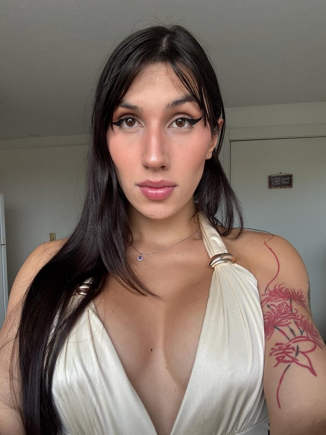A close-up portrait of a person with long dark hair wearing a white tank top, standing indoors near wooden cabinets.