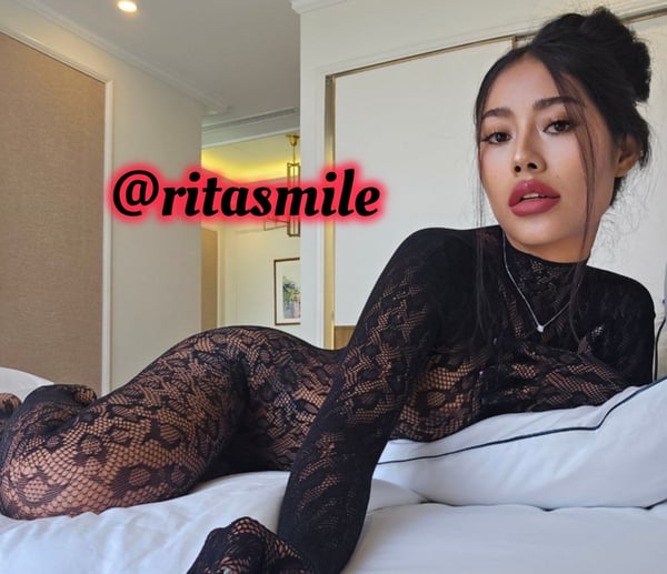 A woman with long dark hair in a bun poses reclined on a bed in a luxurious hotel room with modern decor.