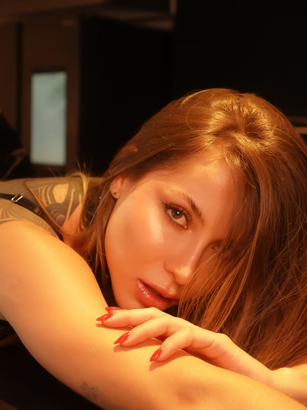 A close-up of a woman with long brown hair and tattoos resting on a surface with warm lighting.