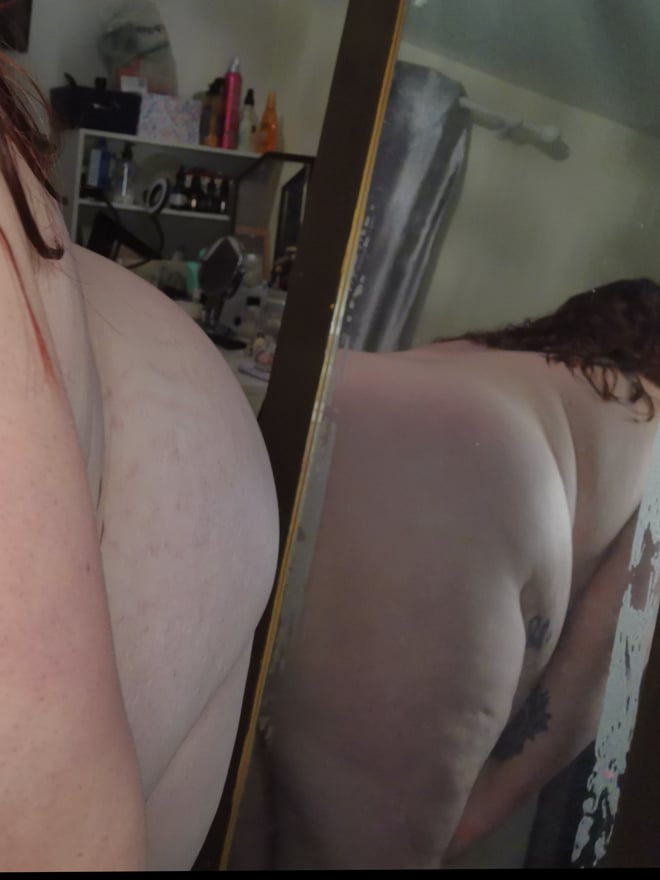 A woman standing in front of a bathroom mirror, viewed from behind, showing her figure.