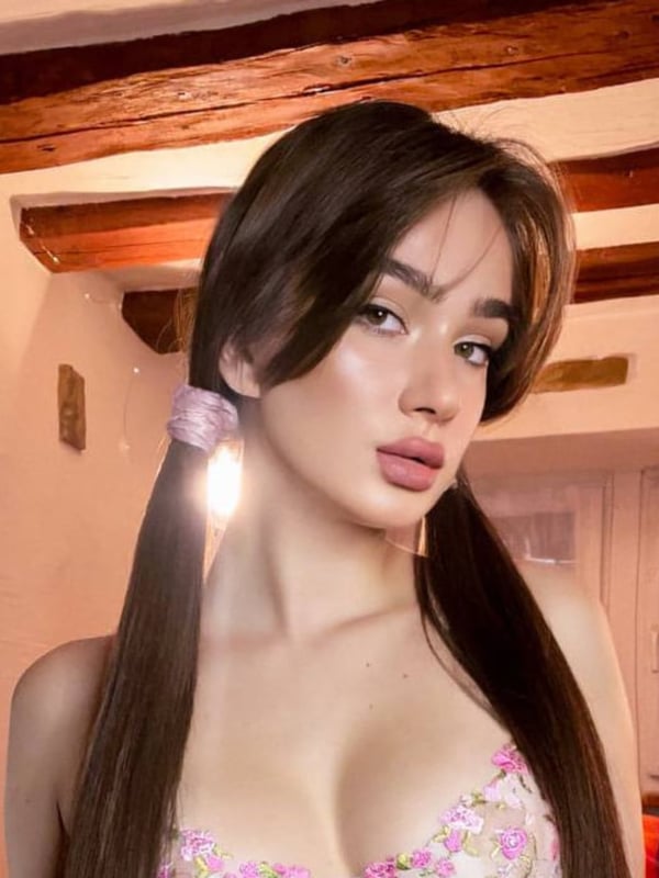 A young woman with long dark hair in pigtails wearing a floral top, posing in a cozy wooden interior.